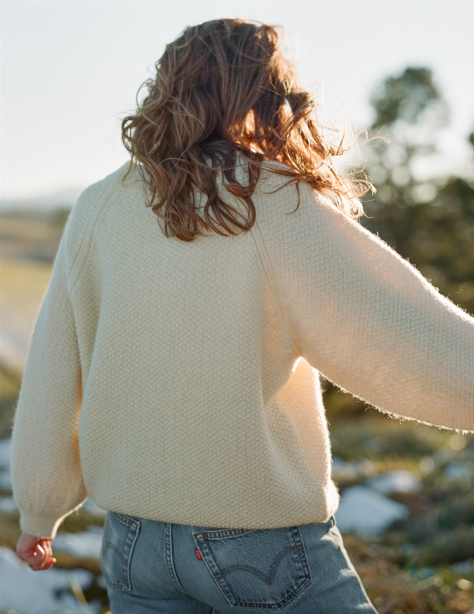 A person with wavy brown hair is seen from the back, wearing a Wol Hide Wool and Alpaca Blend Sweatshirt Sweater in Earth paired with light blue jeans. The individual is outdoors, with blurred greenery and light in the background, suggesting a natural setting.