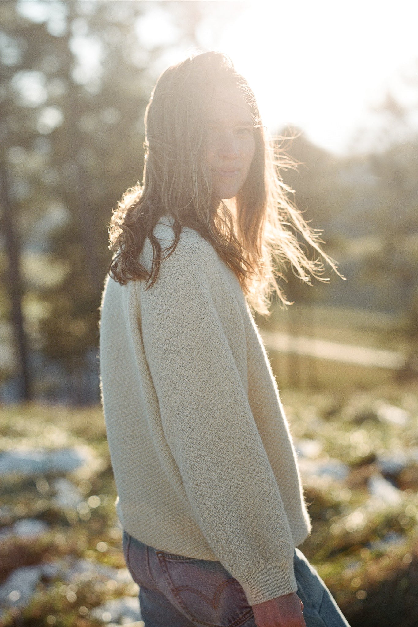 A person with long hair wearing a Wool and Alpaca Blend Sweatshirt Sweater in Natural by Wol Hide is standing outdoors in a sunlit area with blurred trees and grass in the background. The sunlight creates a soft glow around them, and they are looking at the camera with a calm expression.