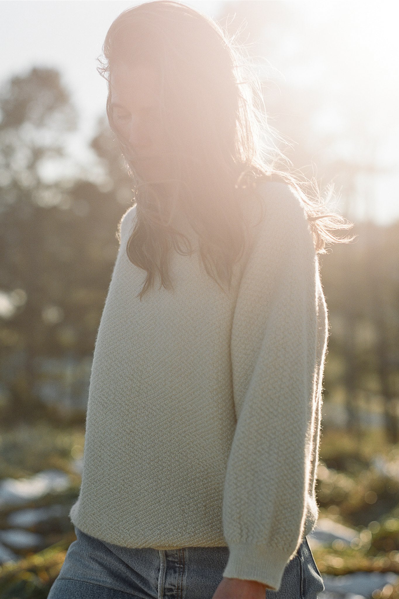 A person with long hair stands outdoors in soft sunlight, wearing the Wol Hide Wool and Alpaca Blend Sweatshirt Sweater in Earth and jeans. Their serene expression complements the slightly blurred background, hinting at trees and a natural setting.