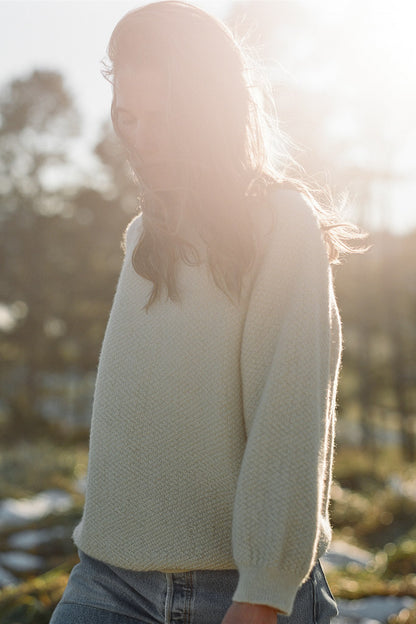 A person with long hair is standing outdoors, bathed in soft sunlight. They are wearing the Wool and Alpaca Blend Sweatshirt Sweater in Natural by Wol Hide, paired with jeans. The background features blurry, sunlit trees, creating a serene and peaceful atmosphere.