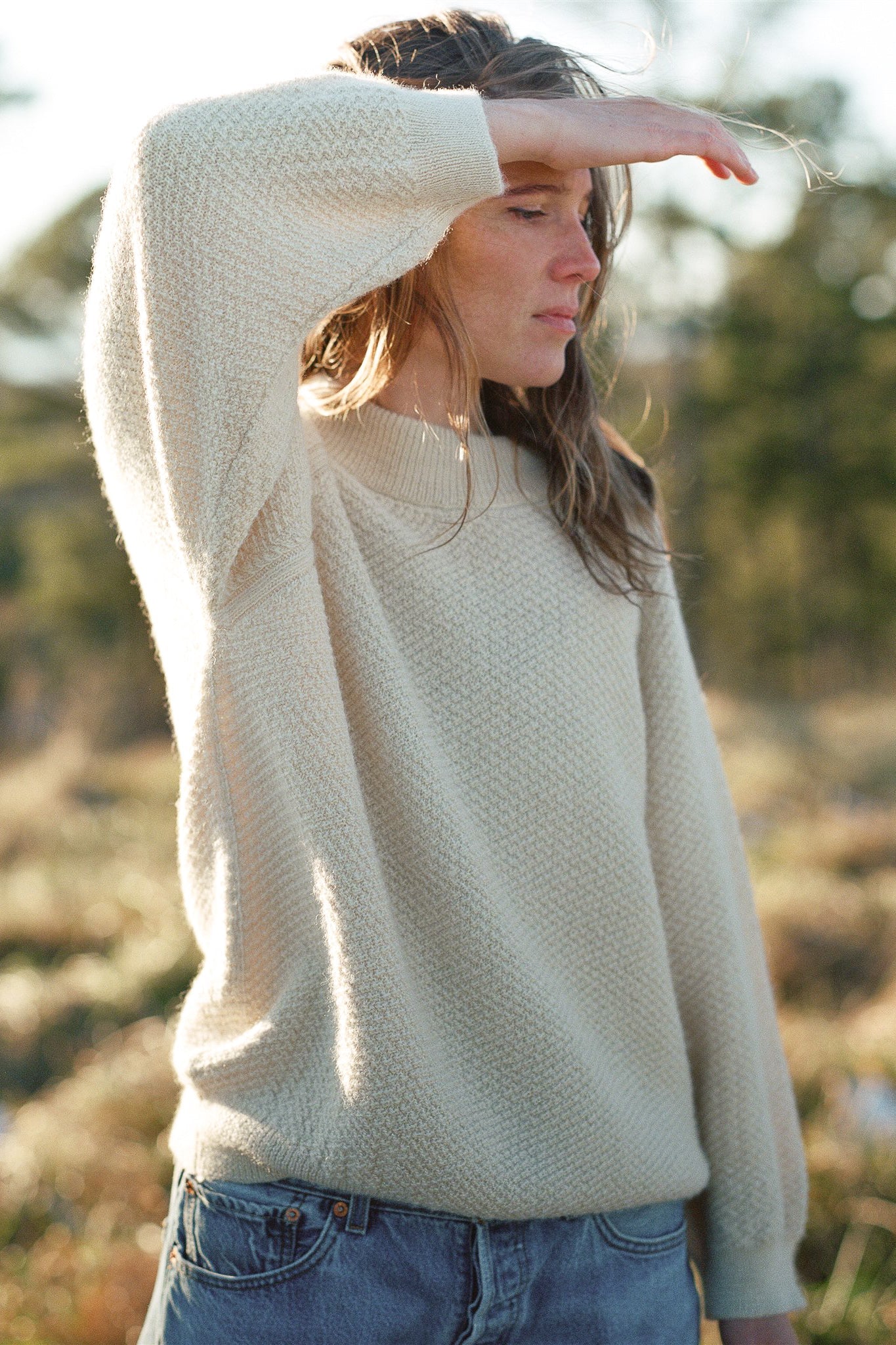 A person with long hair wearing the Wool and Alpaca Blend Sweatshirt Sweater in Earth by Wol Hide and jeans stands outdoors, shading their eyes from the sun. The background is slightly blurred, indicating a natural setting with greenery.