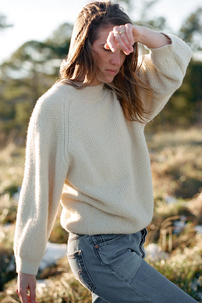 A person with long hair, dressed in a Wool and Alpaca Blend Sweatshirt Sweater in Earth by Wol Hide and grey pants, stands outdoors with their left arm raised, partially covering their face. The background features a blurred view of greenery and trees under bright daylight.