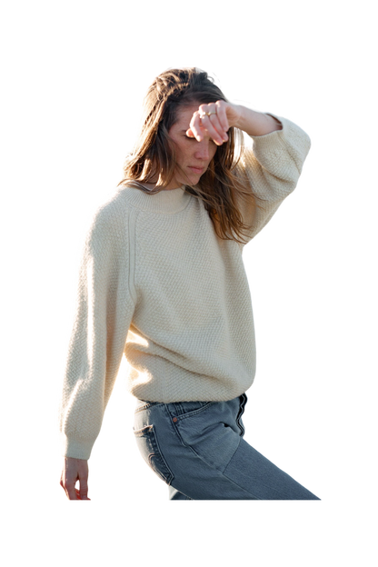 A person with long hair, dressed in a Wol Hide Wool and Alpaca Blend Sweatshirt Sweater in Natural and jeans, is captured mid-motion with one arm raised and the other relaxed by their side. 