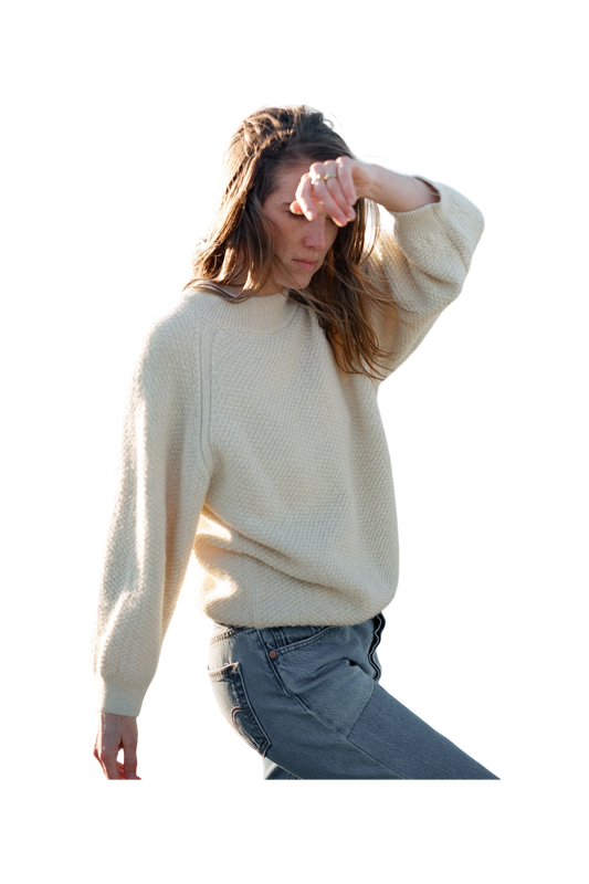 A person with long hair, dressed in a Wol Hide Wool and Alpaca Blend Sweatshirt Sweater in Natural and jeans, is captured mid-motion with one arm raised and the other relaxed by their side. They appear to be outside, and their face is partially obscured by their raised arm.