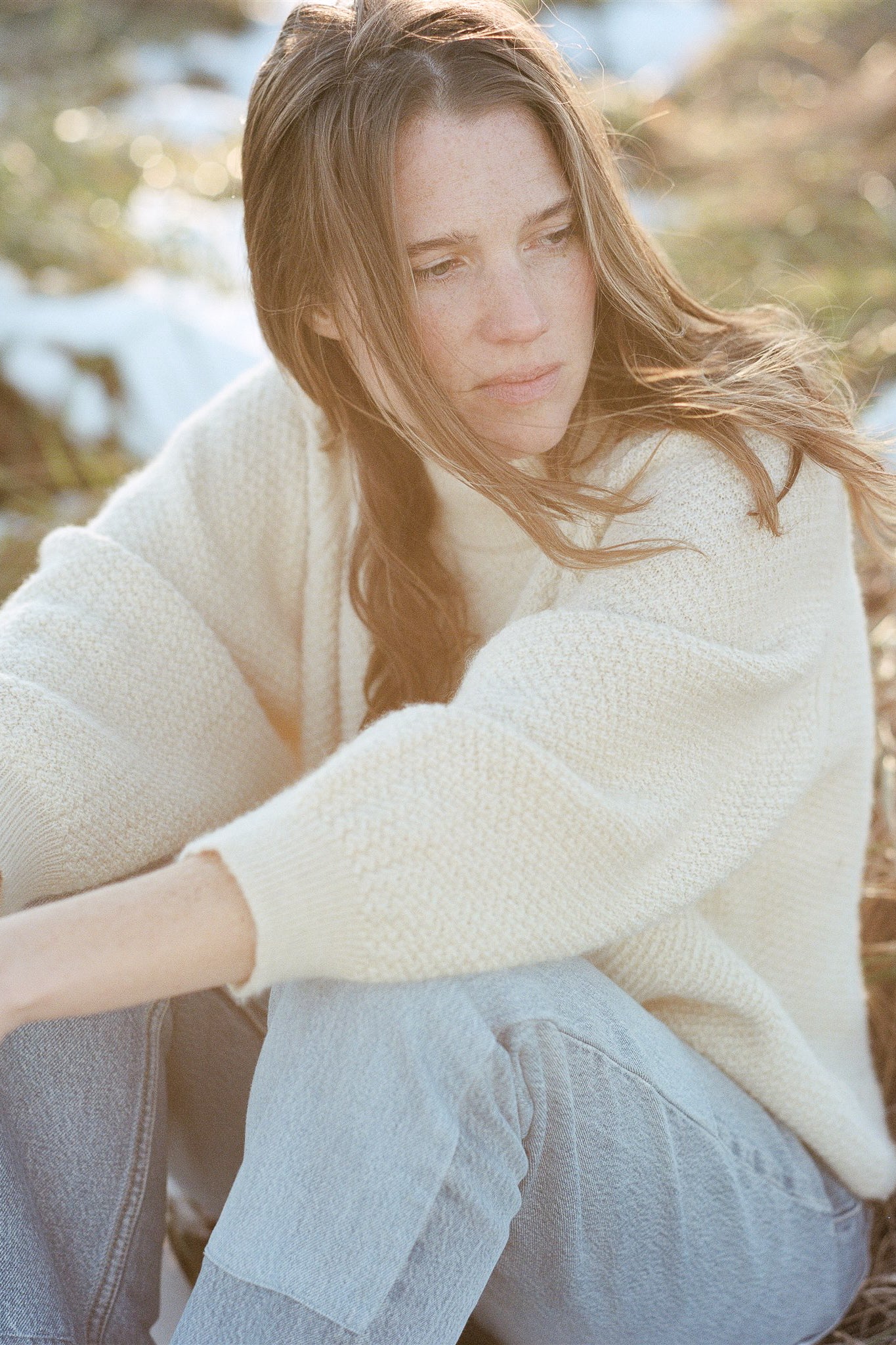 A woman with long hair, wearing a Wol Hide Wool and Alpaca Blend Sweatshirt Sweater in Natural and light blue jeans, sits outdoors on a sunny day. She looks contemplatively into the distance, surrounded by blurred natural scenery, including patches of snow and grass.