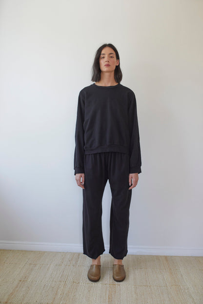 A person with shoulder-length dark hair stands against a plain background. They're wearing a Wol Hide Cotton French Terry Easy Summer Sweatshirt in Black, paired with matching pants made from 100% organic cotton French terry. They also have on brown slip-on shoes. The minimalistic setting features a light-colored floor. Everything is made in the USA.