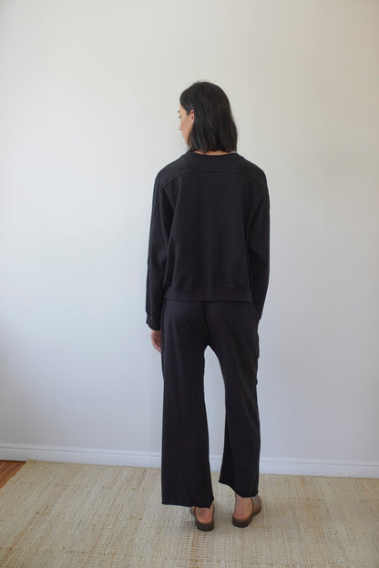 A person with dark hair stands with their back to the camera, wearing a loose-fitting Wol Hide Cotton French Terry Easy Summer Sweatshirt in Black and matching wide-leg pants made from 100% organic cotton French terry. They are standing on a light-colored woven rug against a plain white wall.