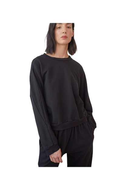 A person with shoulder-length dark hair wearing a loose-fitting Cotton French Terry Easy Summer Sweatshirt in Black by Wol Hide and matching black pants. The individual has their left hand in the pocket of their pants and is looking directly at the camera against a plain background.