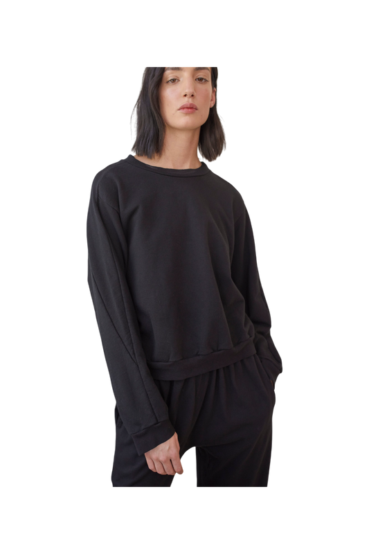 A person with shoulder-length dark hair wearing a loose-fitting Cotton French Terry Easy Summer Sweatshirt in Black by Wol Hide and matching black pants. The individual has their left hand in the pocket of their pants and is looking directly at the camera against a plain background.