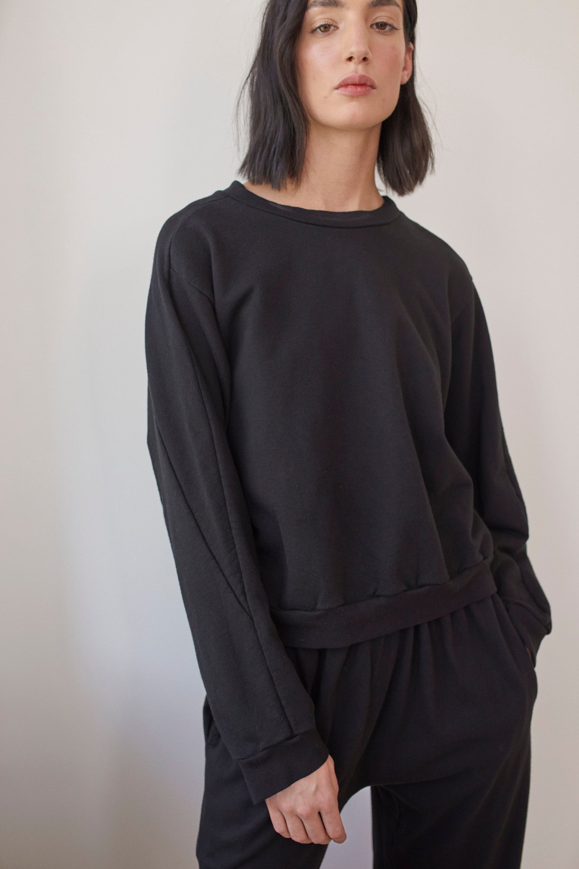 A person with shoulder-length dark hair stands against a plain background, wearing the Wol Hide Cotton French Terry Easy Summer Sweatshirt in black and matching pants made from 100% organic cotton. Their left hand rests in their pocket, and they have a neutral expression on their face.