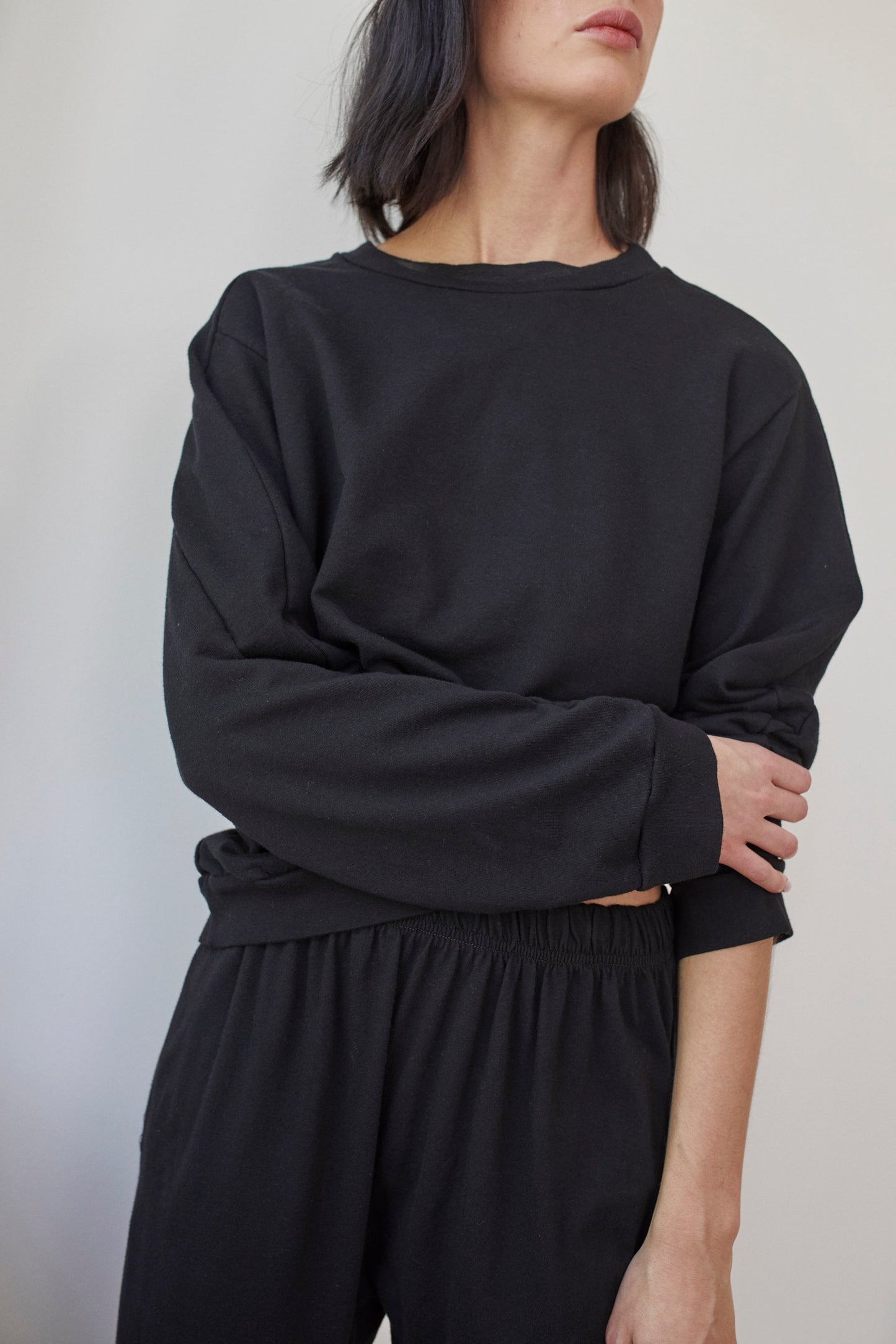 A person with shoulder-length dark hair is wearing Wol Hide’s loose-fitting Cotton French Terry Easy Summer Sweatshirt in black and matching black pants. They are standing against a light-colored background, with one arm crossed over their body and the other hanging down. Their face is partially visible.