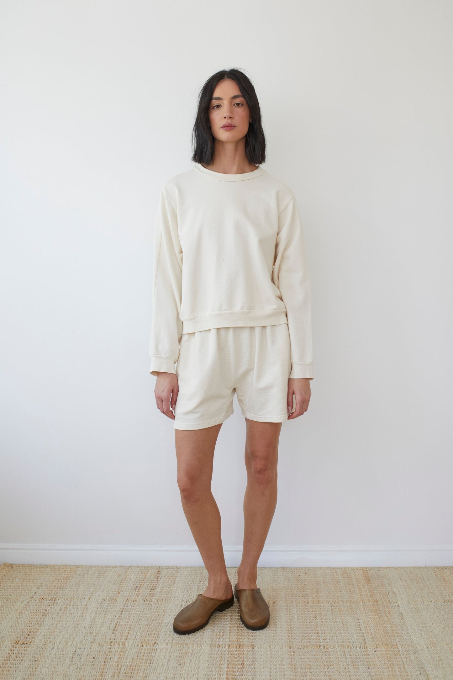 A person with shoulder-length dark hair is standing against a plain white wall. They are wearing an off-white French Terry long-sleeve sweatshirt paired with Wol Hide's Cotton Easy Shorts in Natural and brown slip-on shoes. The floor beneath them is light-colored, and their expression is neutral. Made in the USA.