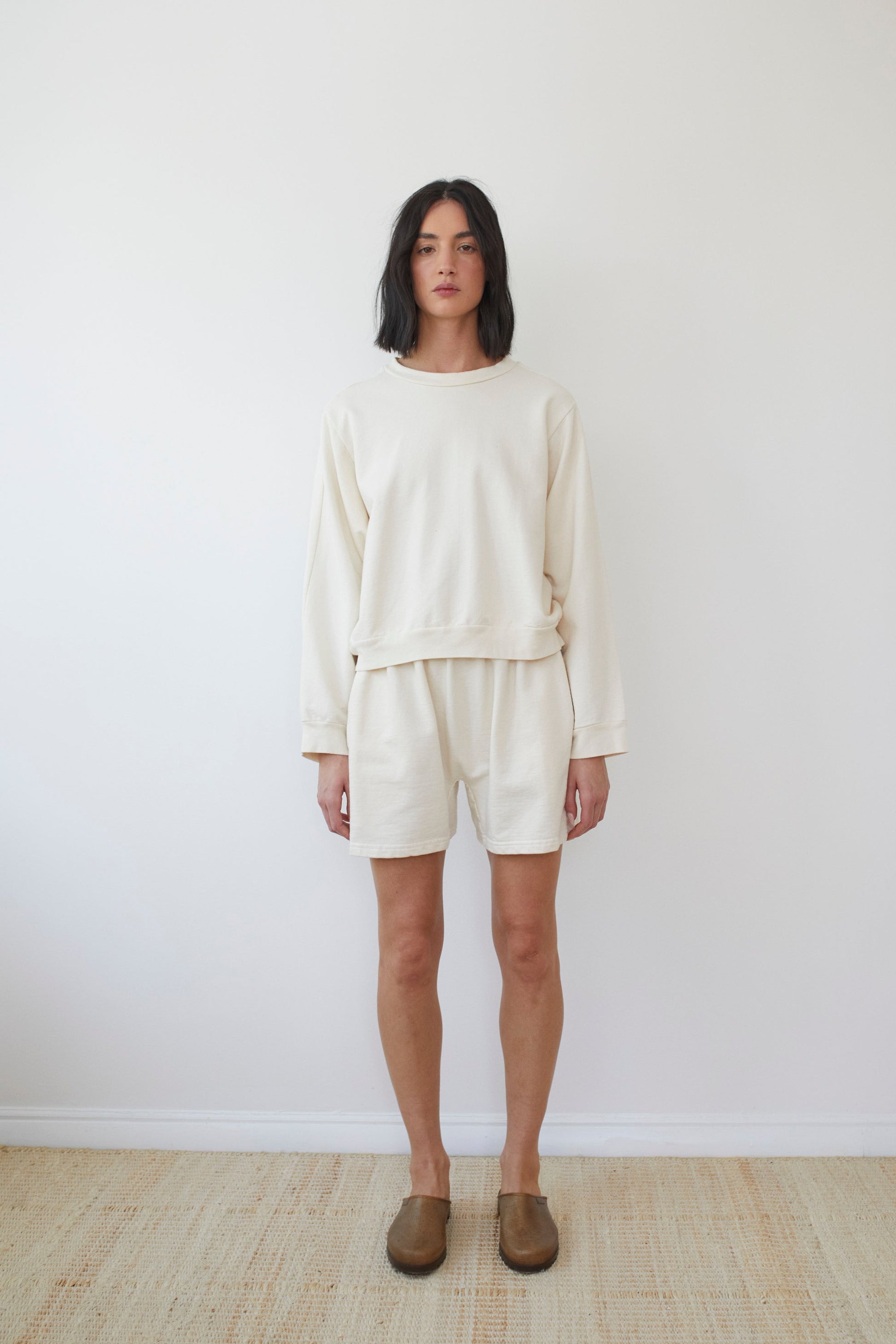 A person with shoulder-length dark hair stands against a plain white wall, wearing the Wol Hide Cotton French Terry Easy Summer Sweatshirt in Natural along with matching shorts made in the USA from organic cotton. They have on brown slip-on shoes and stand on a light-colored woven rug.