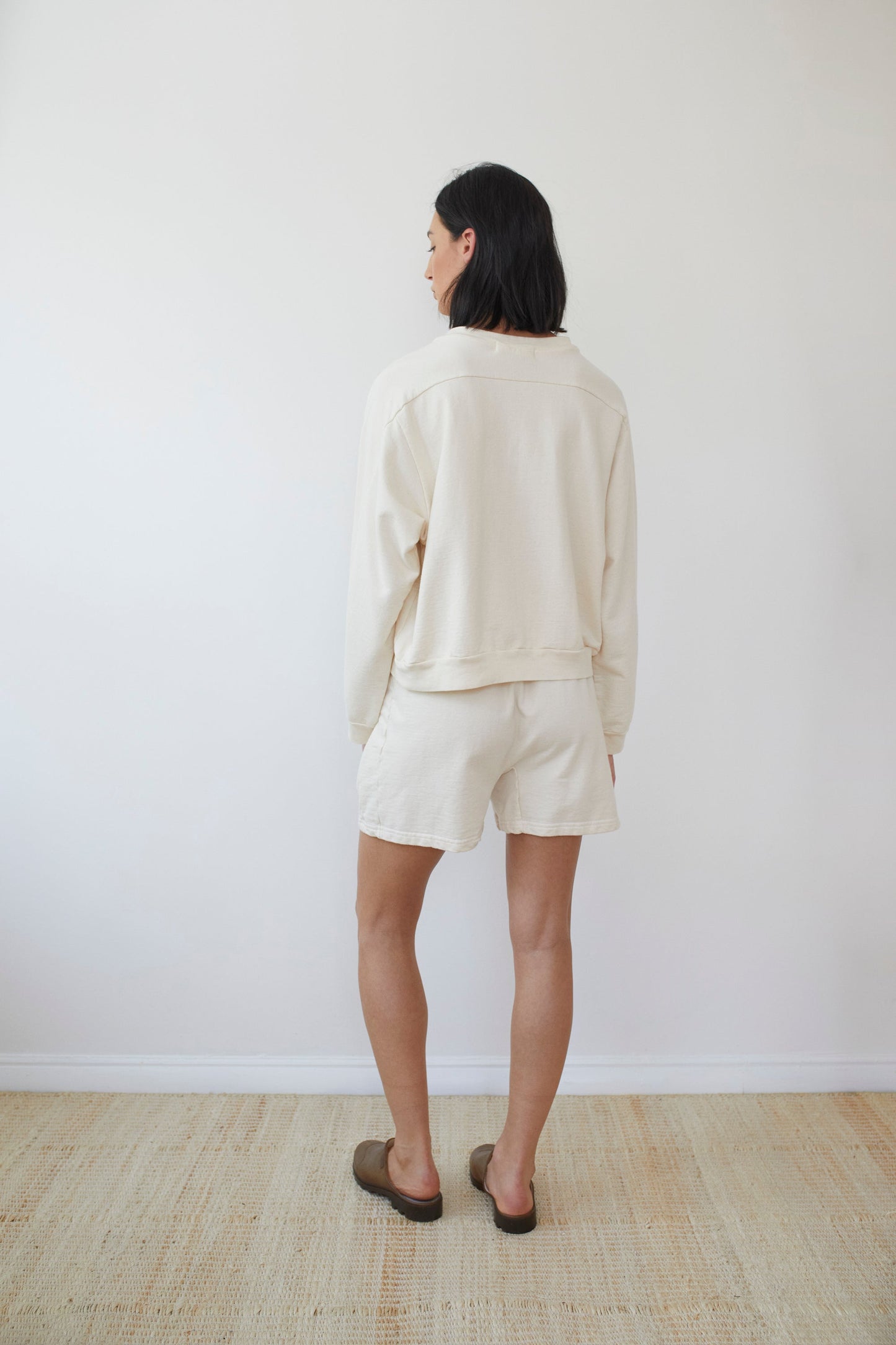 A woman wearing the Wol Hide Organic Cotton French Terry Easy Summer Sweatshirt in Natural and matching shorts stands in a minimalistic room. Her dark shoulder-length hair adds to the scene, complemented by brown slippers, light wooden flooring, and plain white walls.