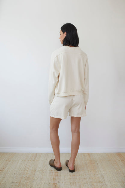 A person with shoulder-length black hair stands facing away from the camera. They are wearing a Cotton French Terry Easy Summer Sweatshirt in Natural by Wol Hide, along with matching shorts and brown slip-on shoes. Crafted from organic cotton and made in the USA, it all stands against a plain white wall and a light-colored woven floor mat.