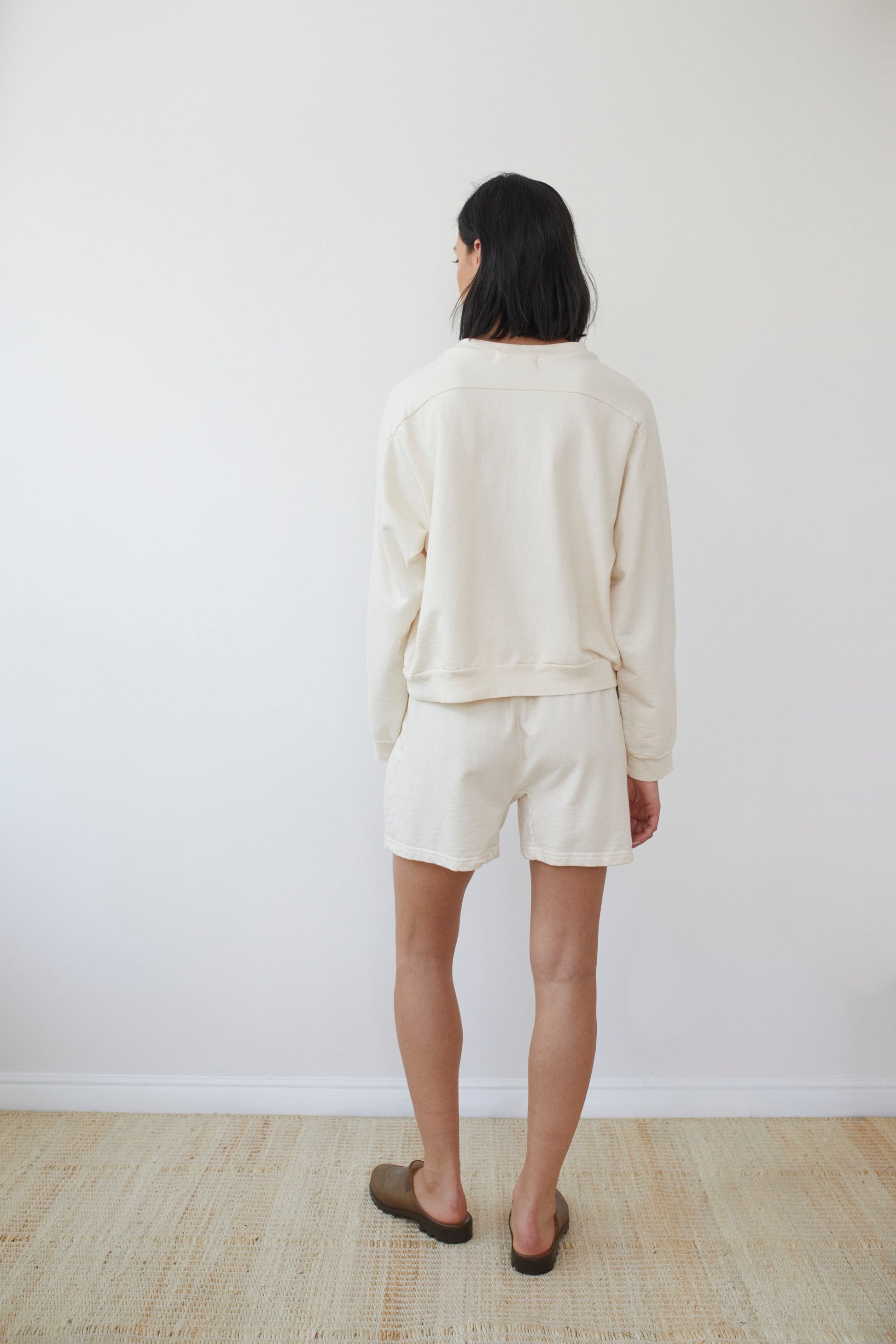 A person with shoulder-length dark hair stands facing away, wearing a cream-colored long-sleeve shirt and Wol Hide's Cotton Easy Shorts in Natural made from organic cotton. They stand on a light-colored woven rug against a plain white wall, sporting brown slip-on shoes.