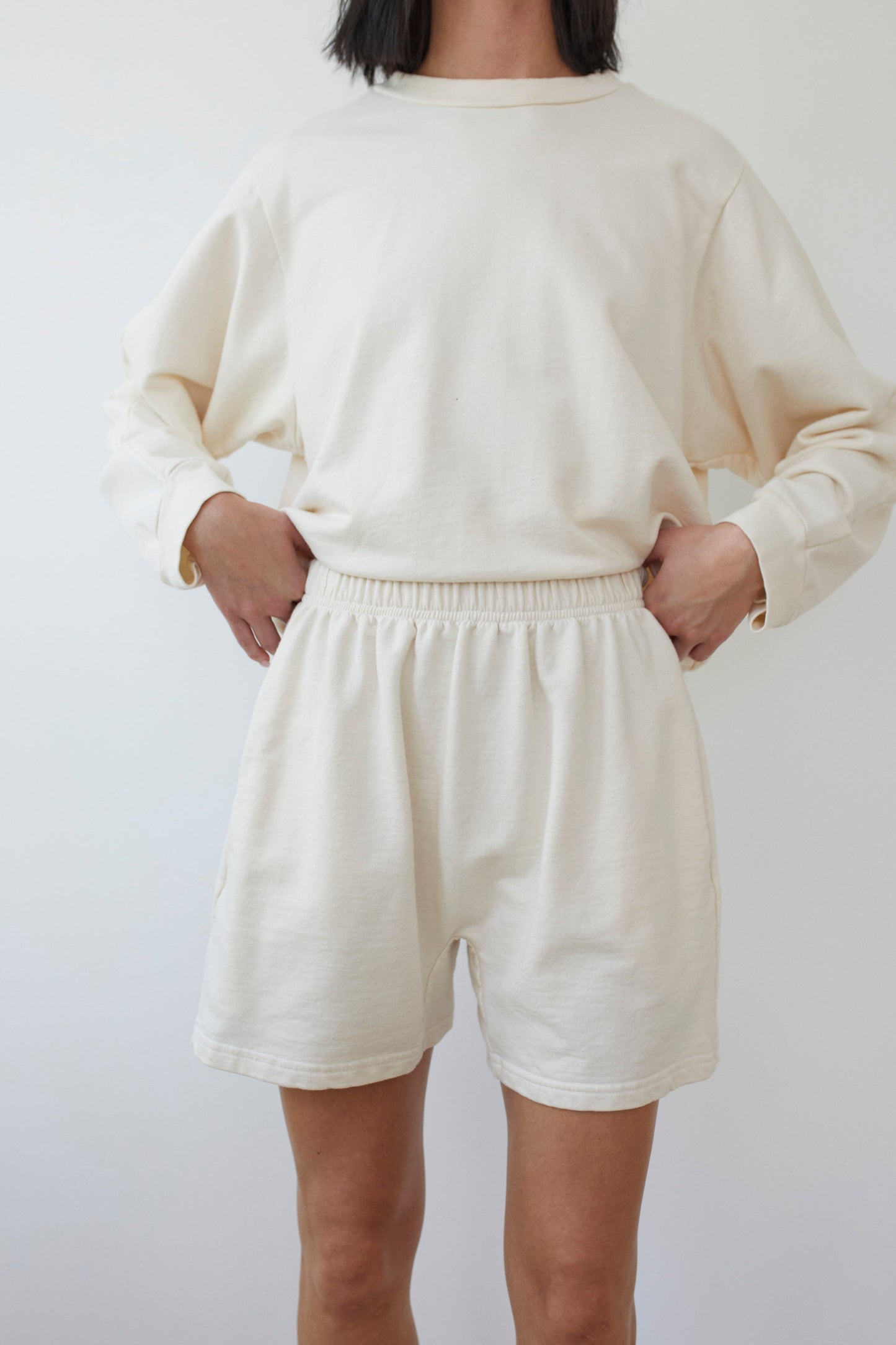 A person standing against a plain, light-colored background is wearing the Cotton Easy Shorts in Natural by Wol Hide, paired with a coordinating cream-colored long-sleeve shirt. The individual has their hands placed on their hips. The outfit, made from soft French Terry fabric, appears casual and comfortable. Made in the USA.