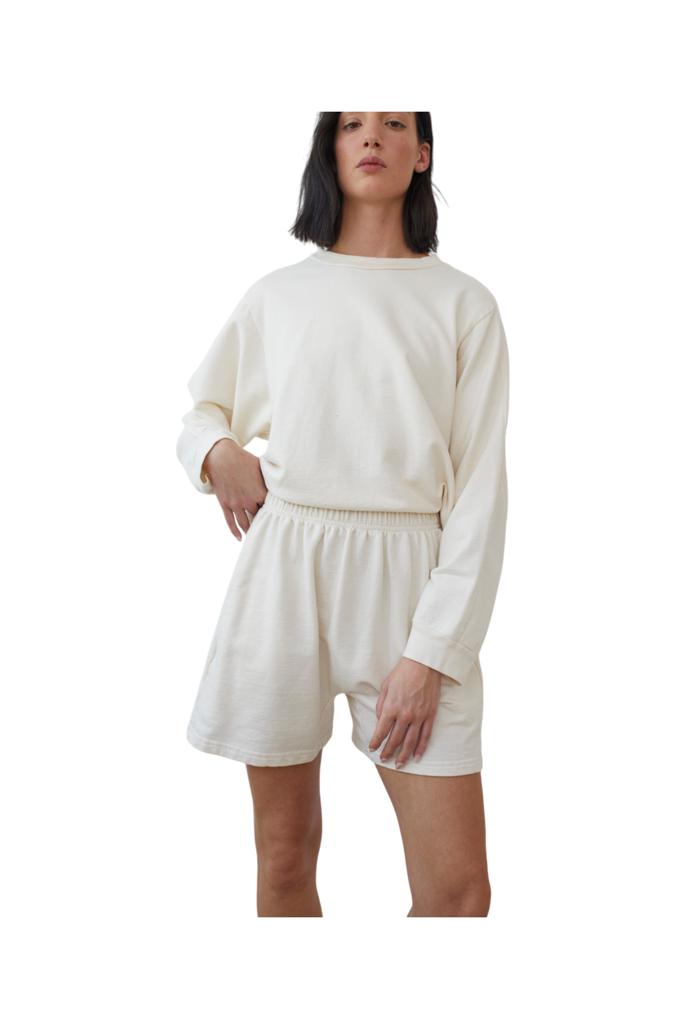 A person with medium-length dark hair is wearing a long-sleeve off-white sweatshirt made of organic cotton and Cotton Easy Shorts in Natural by Wol Hide. They are standing and posing confidently with one hand in their pocket and the other arm relaxed by their side.