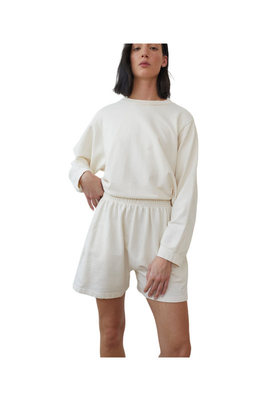 A person with medium-length dark hair is wearing a long-sleeve off-white sweatshirt made of organic cotton and Cotton Easy Shorts in Natural by Wol Hide. They are standing and posing confidently with one hand in their pocket and the other arm relaxed by their side.