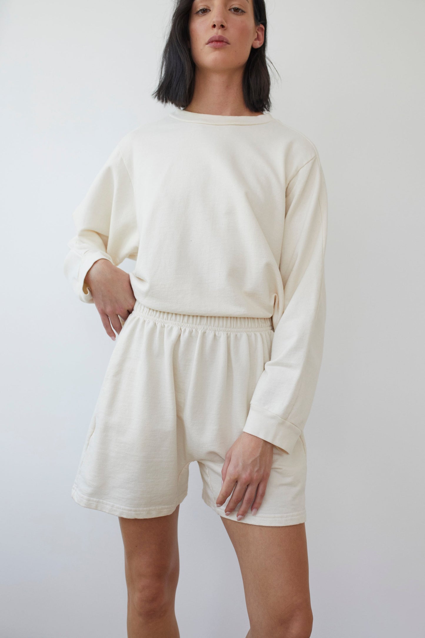 A person with shoulder-length black hair is wearing a loose-fitting, long-sleeved, off-white sweatshirt crafted from organic cotton, paired with Wol Hide's Cotton Easy Shorts in Natural. Made in the USA, the ensemble showcases French Terry fabric. The individual stands against a plain white background, gazing forward with one hand resting on their hip.