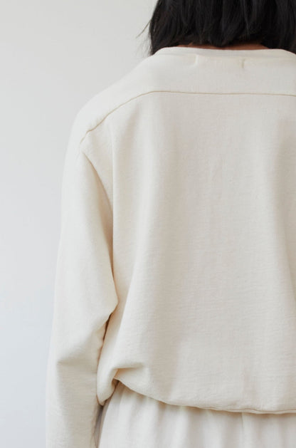 Rear view of a person with long dark hair wearing the Wol Hide Organic Cotton French Terry Easy Summer Sweatshirt in Natural against a plain, light-colored wall.