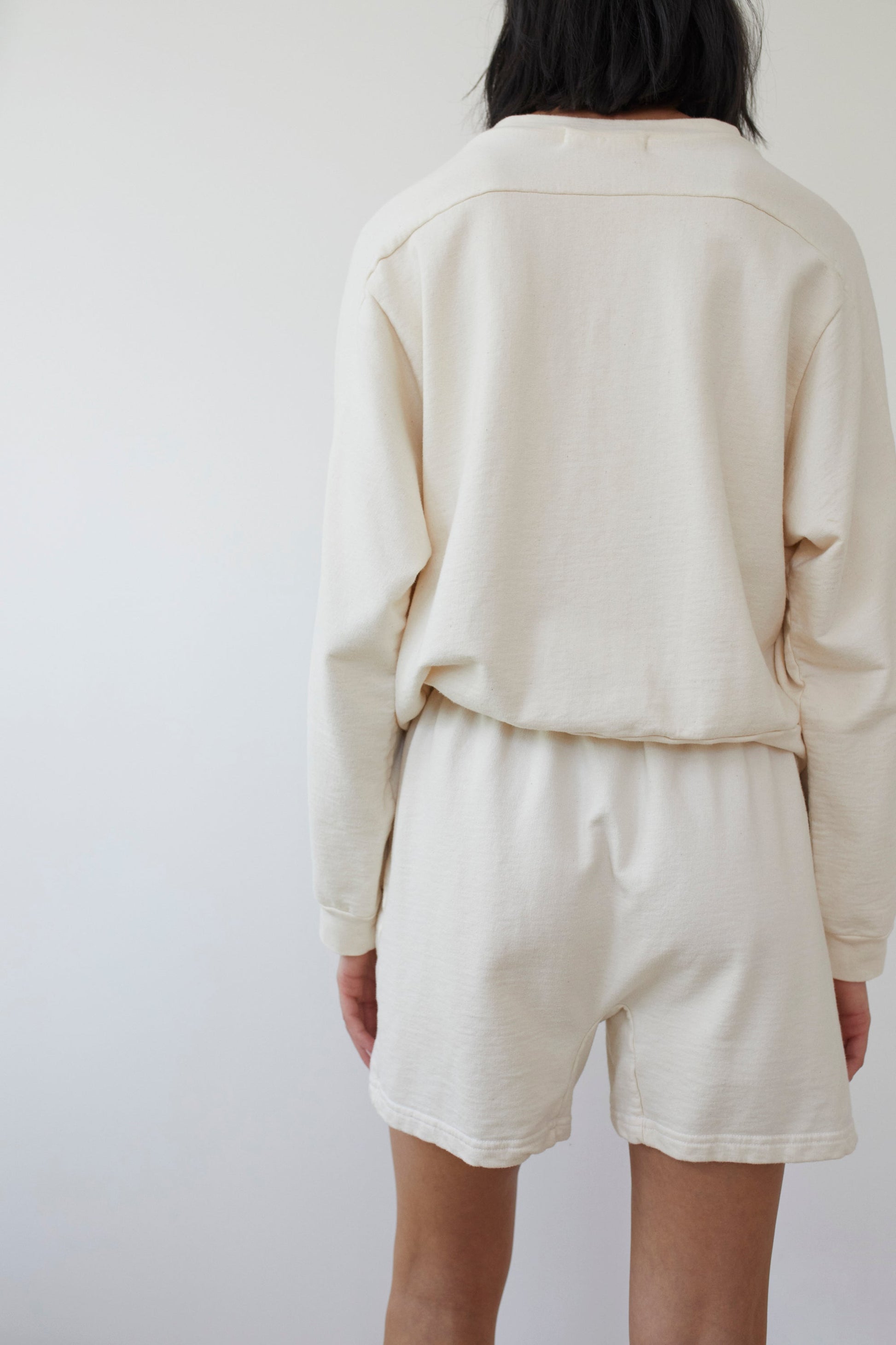 A person with shoulder-length dark hair stands with their back to the camera, wearing a light beige long-sleeve top and Wol Hide's Cotton Easy Shorts in Natural, made of organic cotton. The background is plain and light-colored.