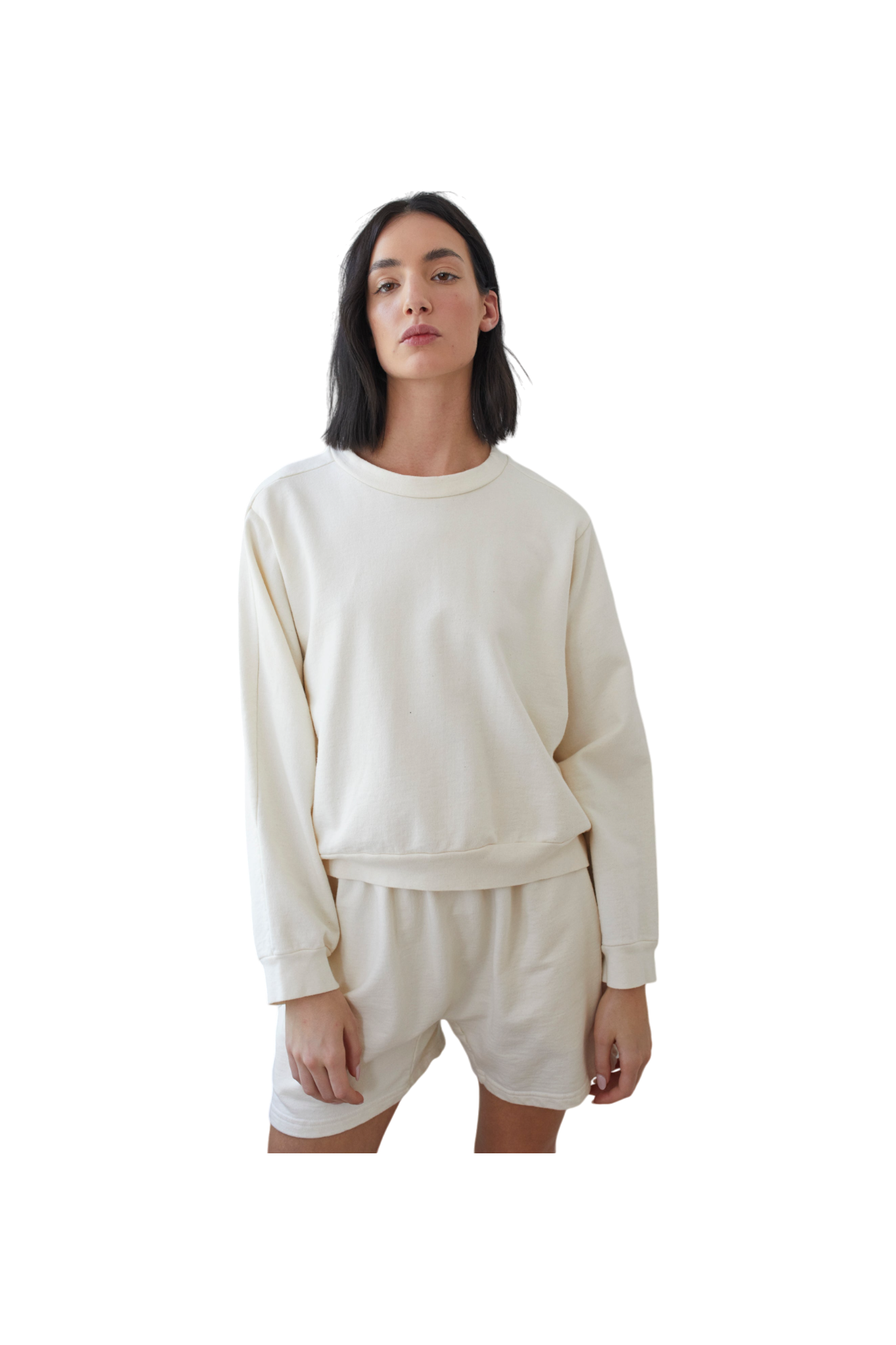 A person with long, dark hair wears Wol Hide's Organic Cotton French Terry Easy Summer Sweatshirt in Natural and matching shorts against a plain, dark background, looking straight ahead with a neutral expression.