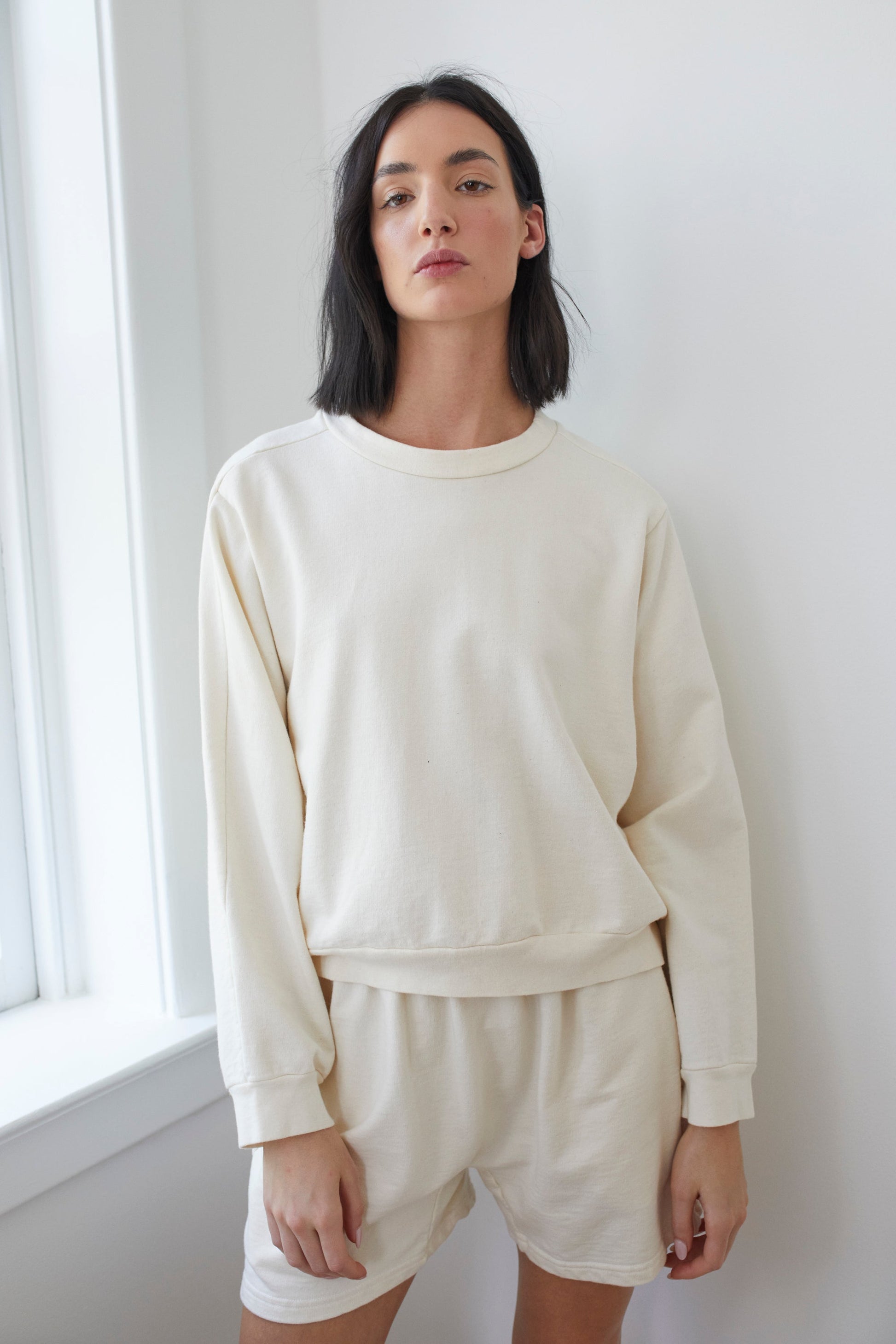 With shoulder-length black hair, someone wears the Wol Hide Organic Cotton French Terry Easy Summer Sweatshirt in Natural and matching shorts, crafted in the USA. They stand relaxed by a white wall near a window, their expression neutral.