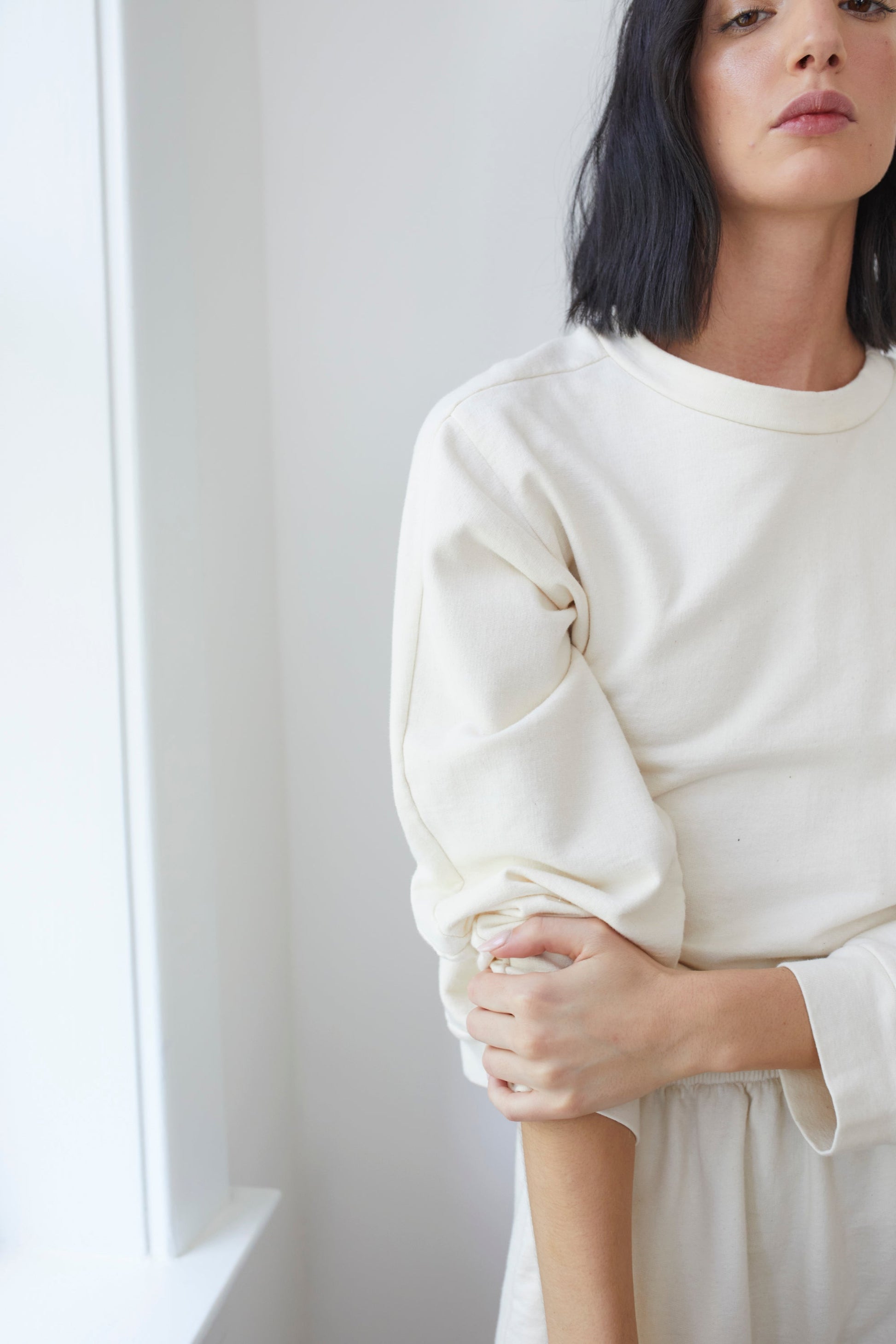 A person with shoulder-length dark hair and light skin stands by a white wall, wearing the Cotton French Terry Easy Summer Sweatshirt in Natural by Wol Hide. One arm is bent, and the hand of the other arm is gently holding the bent arm's elbow. The background is softly lit, creating a calm atmosphere.