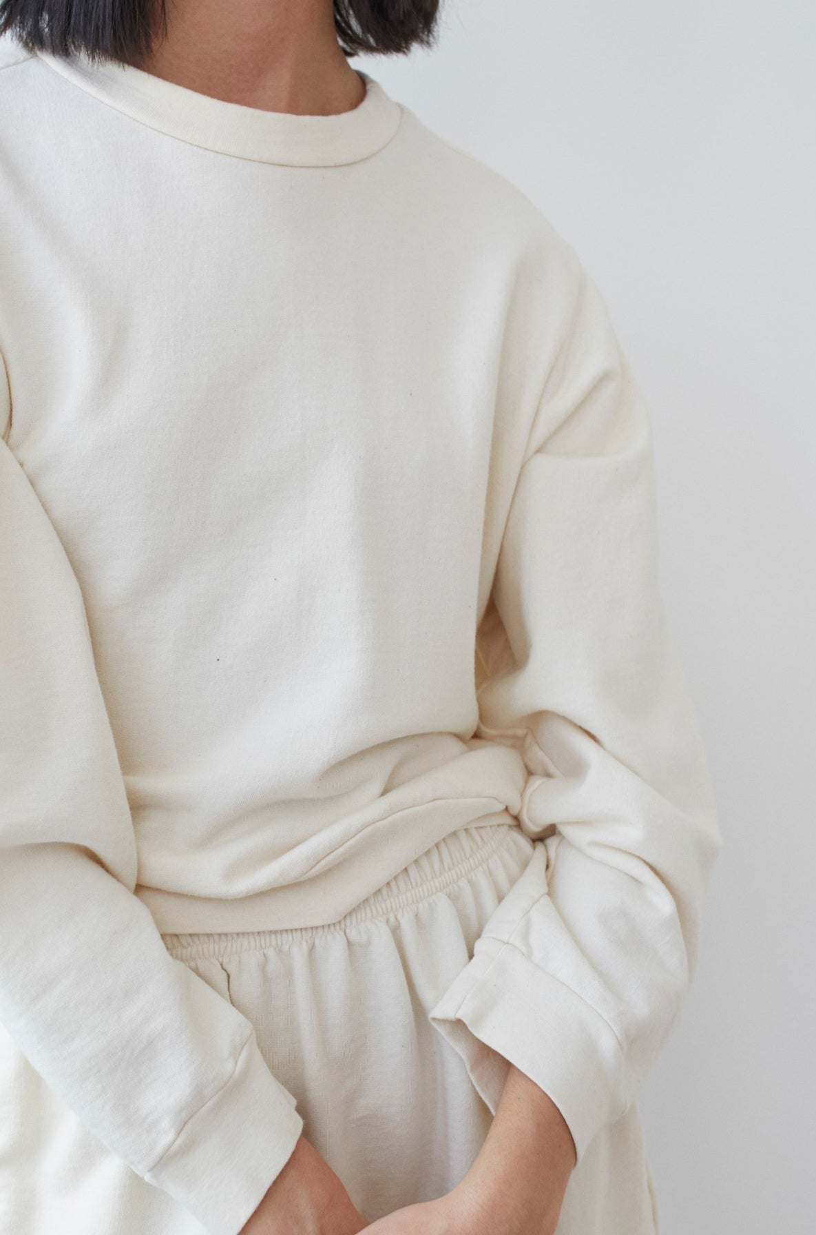 A person with short, dark hair is shown from the shoulders down, wearing the Wol Hide Cotton French Terry Easy Summer Sweatshirt in Natural along with matching pants made of organic cotton French terry. The outfit appears to be comfortable and casual. The background is a plain, light-colored wall.