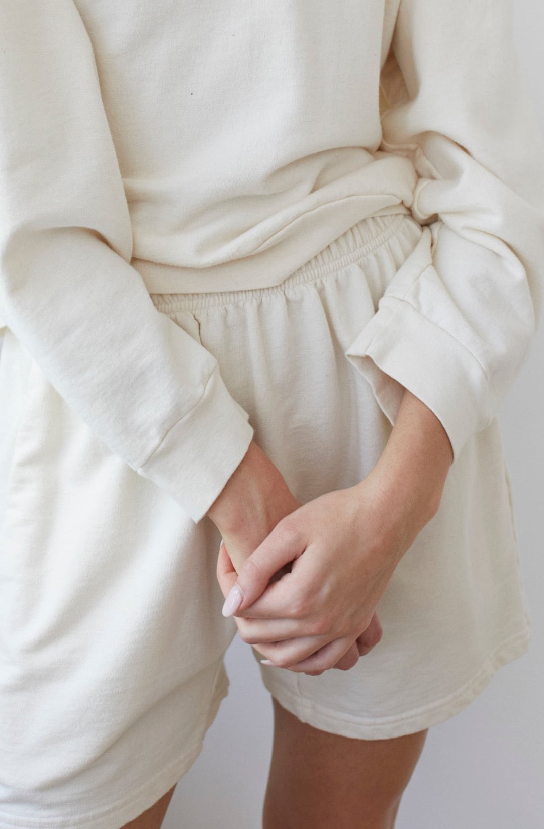 A person wearing a matching light beige French Terry sweater and Wol Hide's Cotton Easy Shorts in Natural stands with their hands clasped in front of them. The outfit, made in the USA, is loose and comfortable, and the person's nails are clean and neatly trimmed. The background is blurred and light-colored.