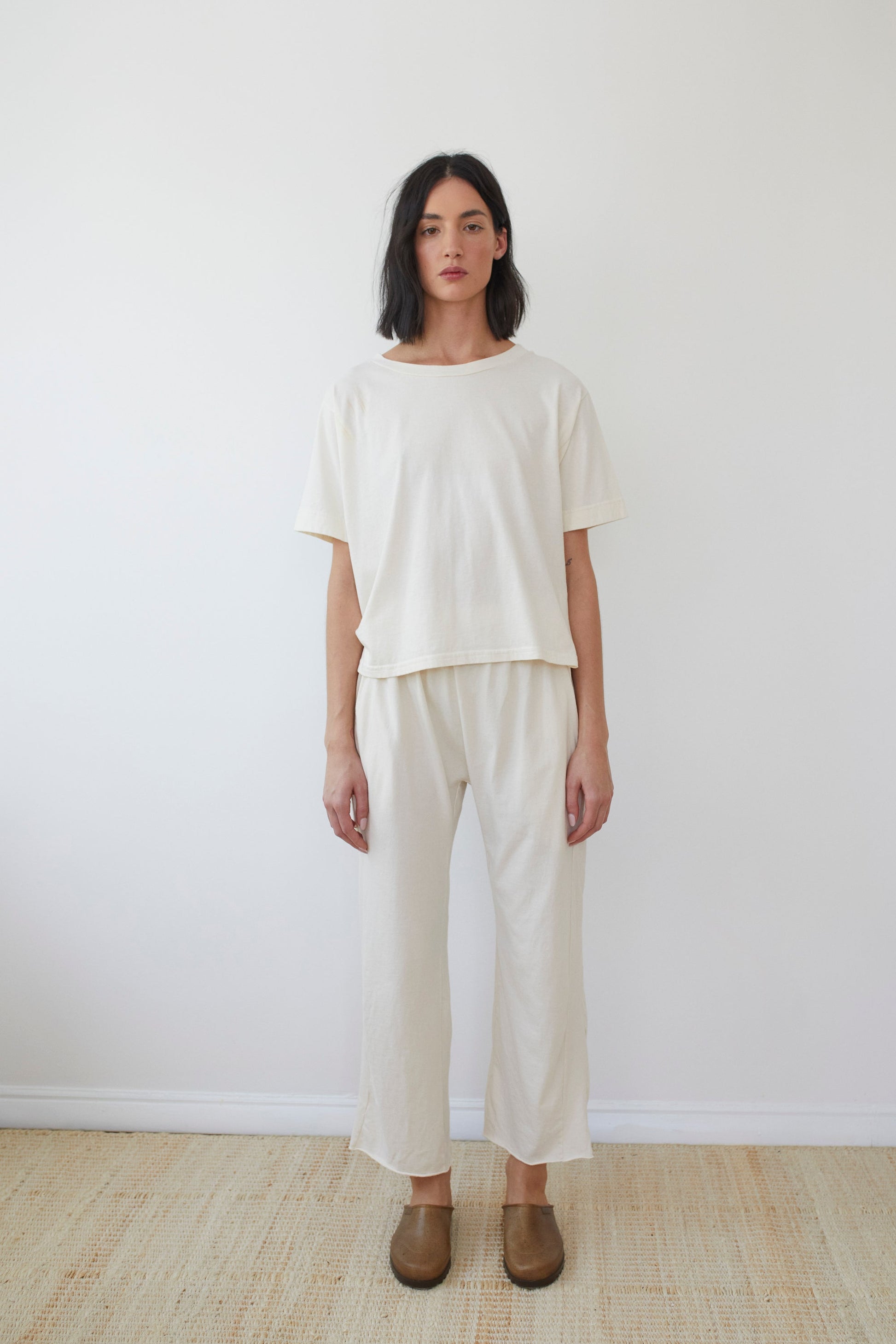 A person with shoulder-length dark hair stands against a plain white background, wearing the Wol Hide Cotton Jersey Box Tee in Natural made from organic cotton and matching loose-fitting pants. They are also wearing brown slip-on shoes and standing on a light-colored woven mat. Made in the USA.