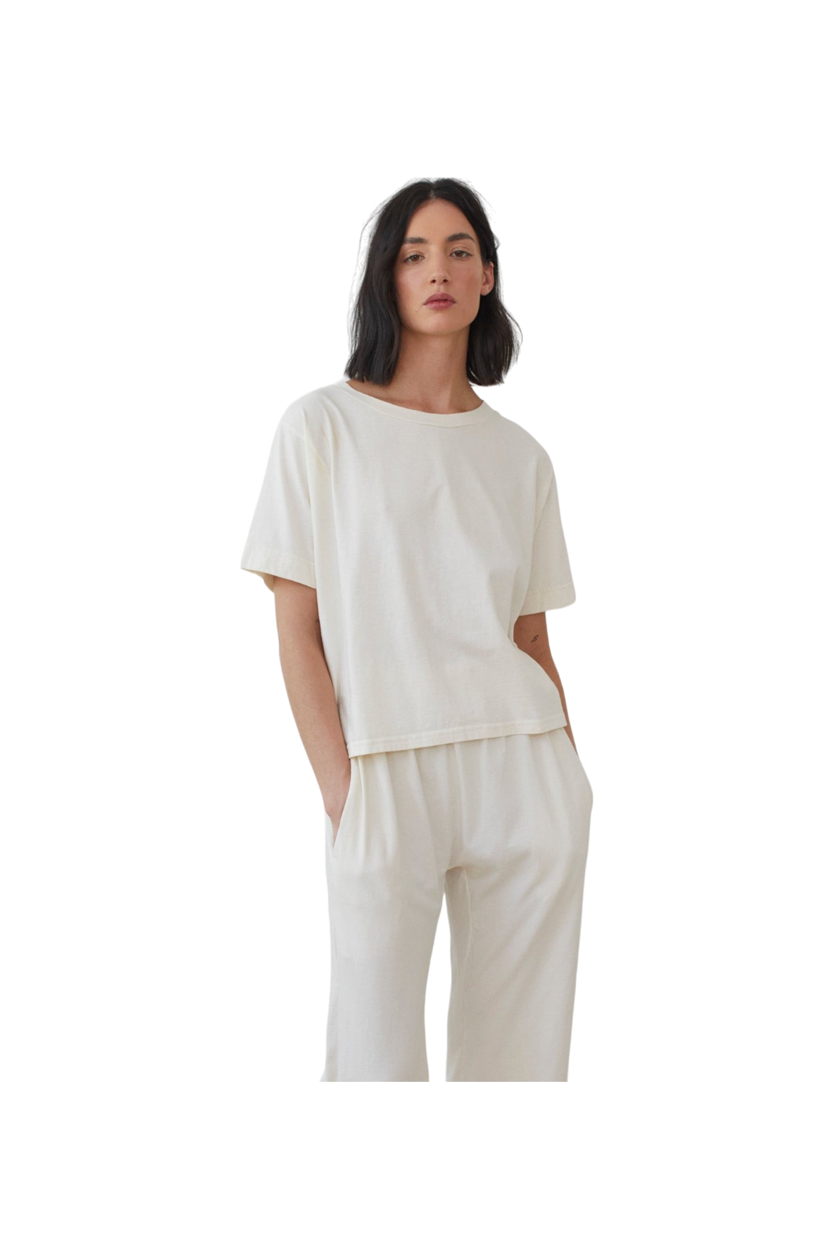 A person with shoulder-length black hair is wearing the Wol Hide Cotton Jersey Box Tee in Natural, paired with matching white pants made from lightweight jersey. The person has their hands in their pockets and is looking slightly to the side against a plain white background.