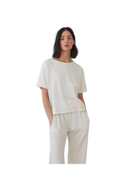 A person with shoulder-length black hair is wearing the Wol Hide Cotton Jersey Box Tee in Natural, paired with matching white pants made from lightweight jersey. The person has their hands in their pockets and is looking slightly to the side against a plain white background.