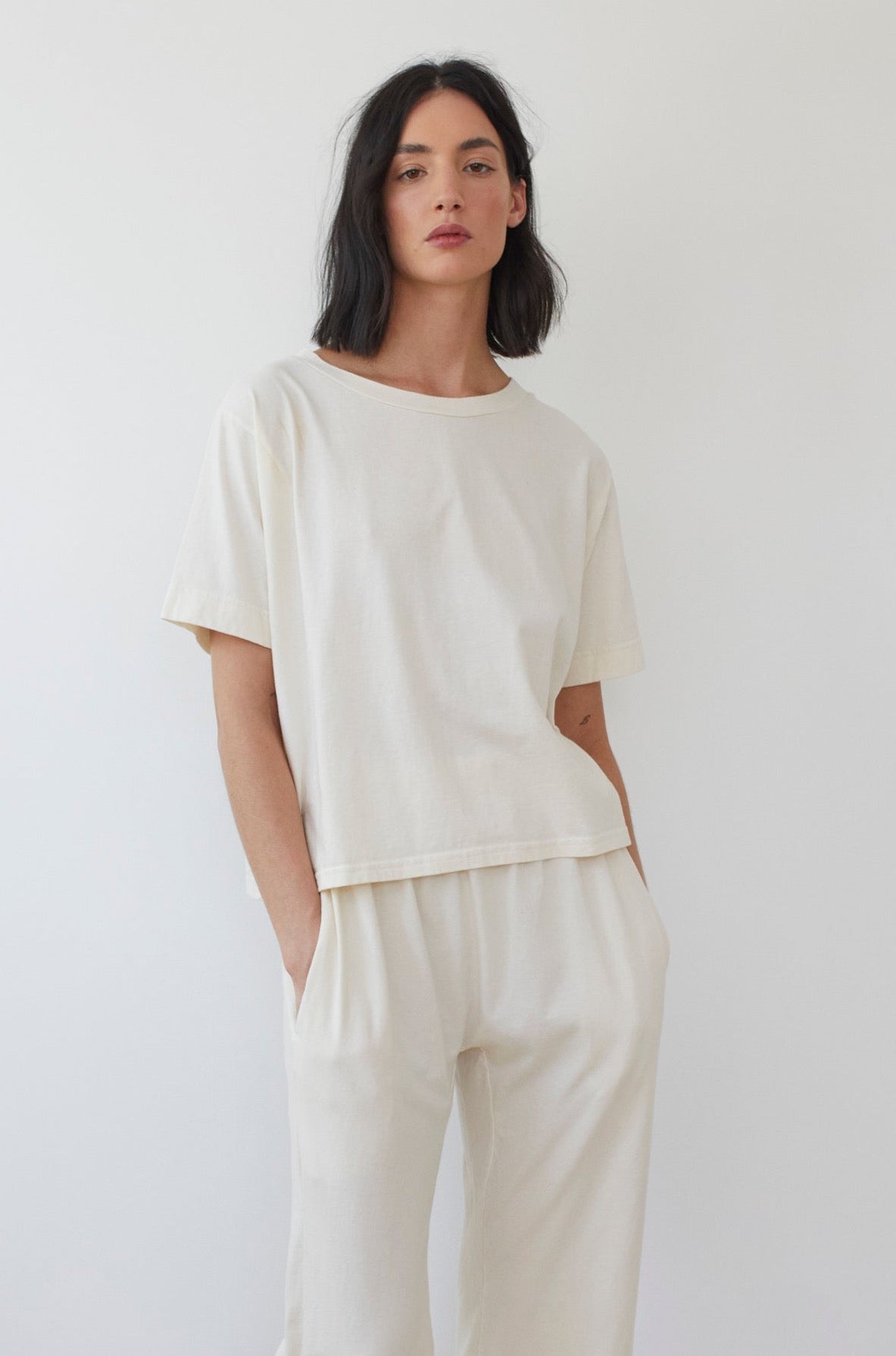 A person with shoulder-length dark hair stands against a plain background, wearing a loose-fitting Cotton Jersey Box Tee in Natural by Wol Hide, along with matching wide-leg organic cotton pants. The person has one hand in their pocket and maintains a neutral expression.