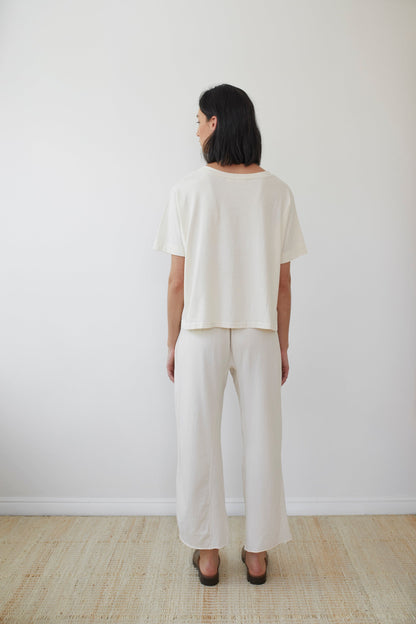 A person with shoulder-length dark hair stands facing away from the camera against a plain white wall. They are wearing Wol Hide's loose-fitting, short-sleeved Cotton Jersey Box Tee in Natural and matching wide-leg pants made from lightweight jersey organic cotton. Their hands are relaxed by their sides as they stand on a beige woven rug.