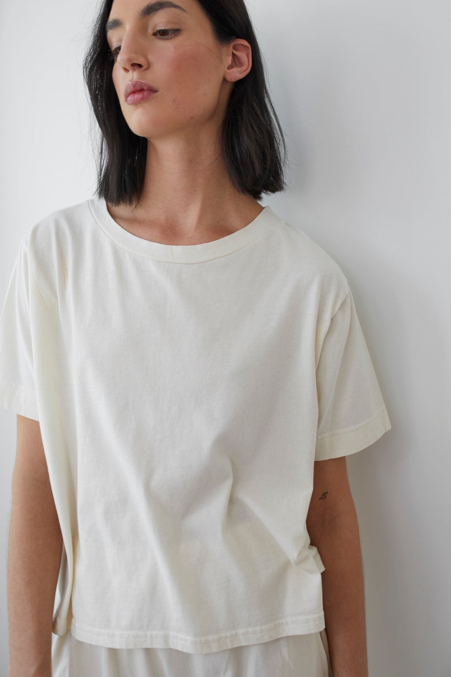 A person with shoulder-length dark hair is wearing the Wol Hide Cotton Jersey Box Tee in Natural, which features short sleeves and is made of lightweight jersey. They are standing against a white wall, gazing slightly to the side. The overall look is minimal and relaxed.