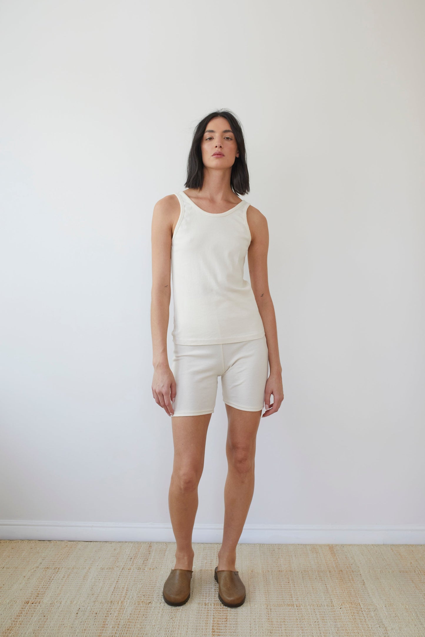 A person stands against a plain white background wearing Wol Hide's sleeveless top and Organic Cotton Bike Shorts in Natural, both made in the USA. They have short dark hair and sport brown slip-on shoes on a light-colored floor, enhancing the clean, minimalist aesthetic.