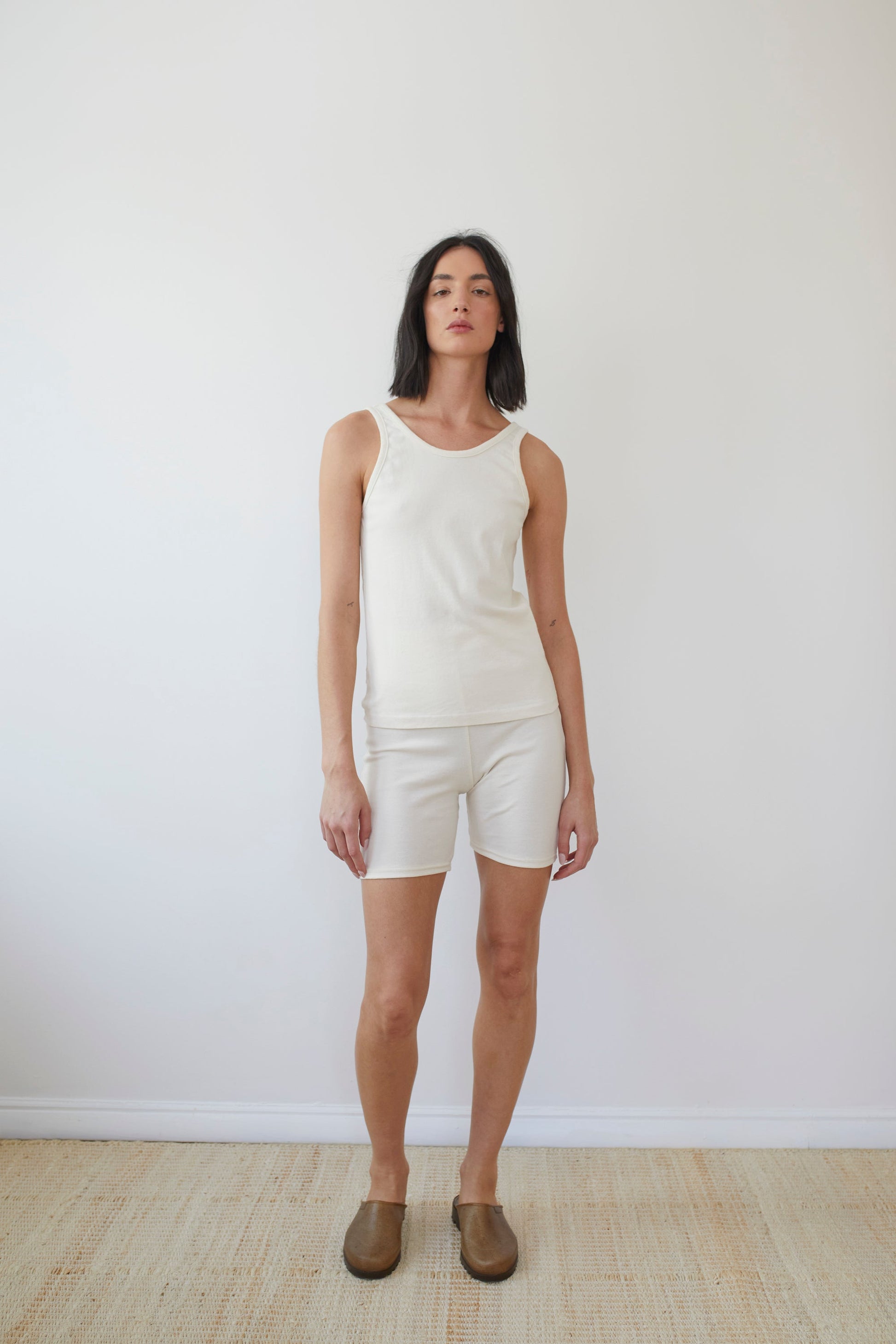 A person with shoulder-length dark hair stands in front of a plain white wall. They are wearing a white sleeveless top and Wol Hide's Cotton Bike Shorts in Natural, paired with light brown slip-on shoes. The floor is a light beige color. The overall scene is minimalist and simple, embodying sustainable clothing choices.
