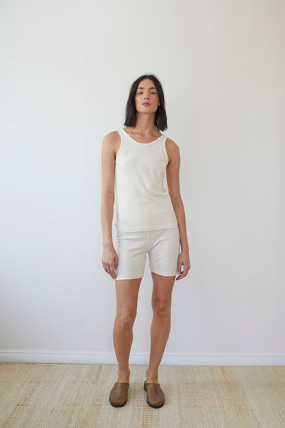 A person stands against a plain white background wearing Wol Hide's sleeveless top and Organic Cotton Bike Shorts in Natural, both made in the USA. They have short dark hair and sport brown slip-on shoes on a light-colored floor, enhancing the clean, minimalist aesthetic.