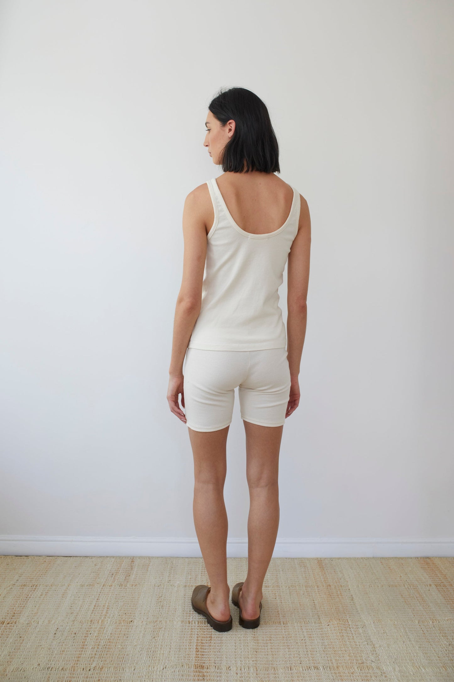 A person with short dark hair stands facing away from the camera, wearing a sleeveless white top and Wol Hide's Cotton Bike Shorts in Natural. They are standing on a light-colored woven rug against a plain white wall. They are also wearing brown slip-on shoes.