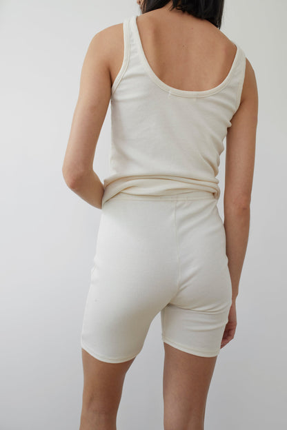 A person with short dark hair is facing away from the camera, wearing a white tank top and Wol Hide's Cotton Bike Shorts in Natural. Made in the USA, this sustainable clothing choice stands out against the plain, white background.