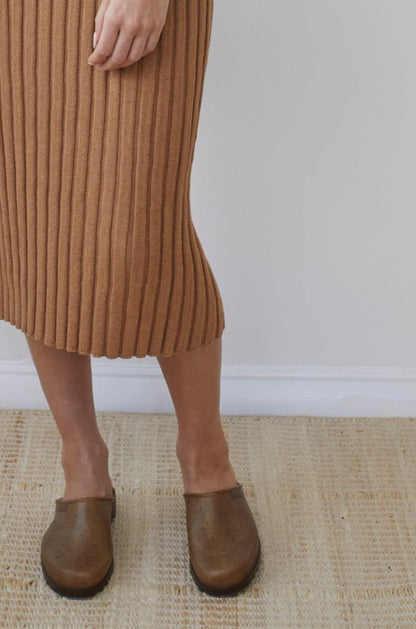 A person standing on a light-colored textured rug, wearing the Wol Hide Rib Dress in Toffee made from organic regenerative cotton and brown clogs. The upper body is not visible.