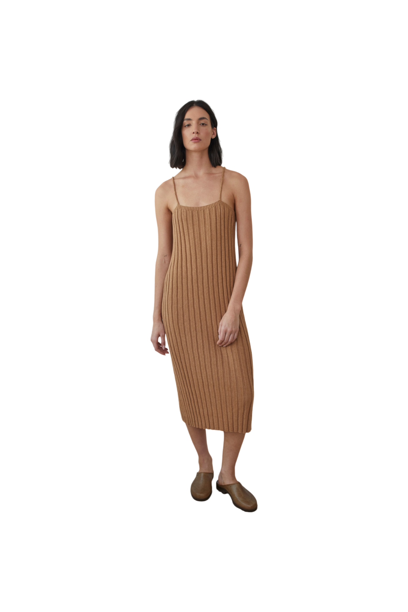 A woman stands against a black background, wearing the Rib Dress in Toffee by Wol Hide, complemented by brown slip-on shoes. Her shoulder-length dark hair frames her look.