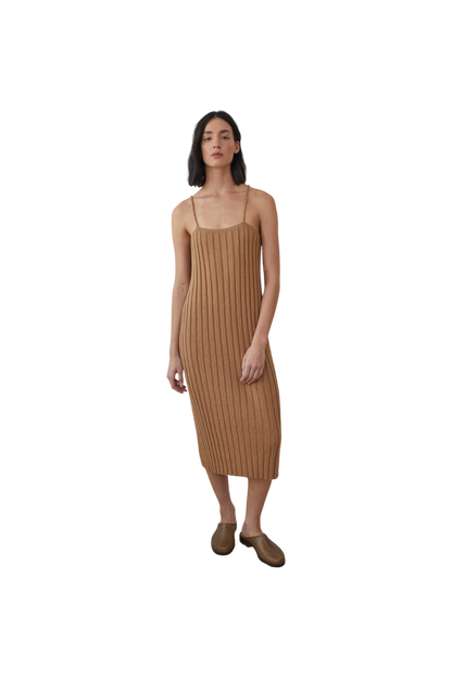 A woman stands against a black background, wearing the Rib Dress in Toffee by Wol Hide, complemented by brown slip-on shoes. Her shoulder-length dark hair frames her look.