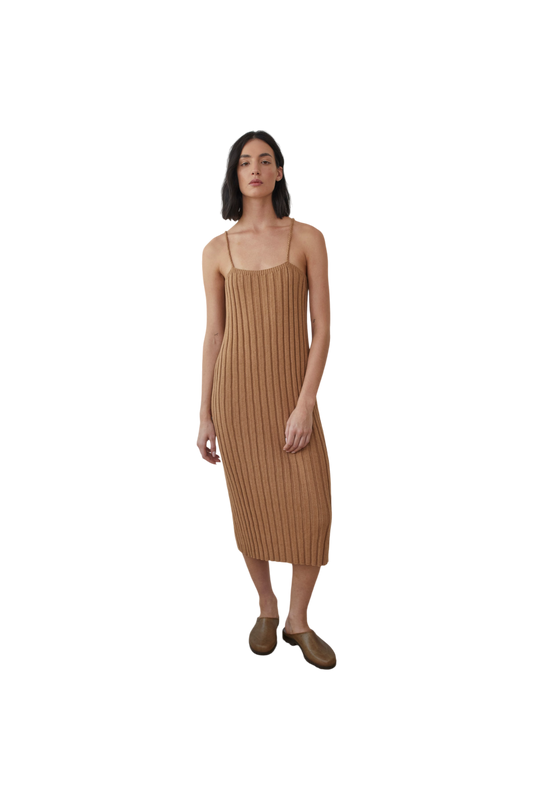 A woman stands against a black background, wearing the Rib Dress in Toffee by Wol Hide, complemented by brown slip-on shoes. Her shoulder-length dark hair frames her look.