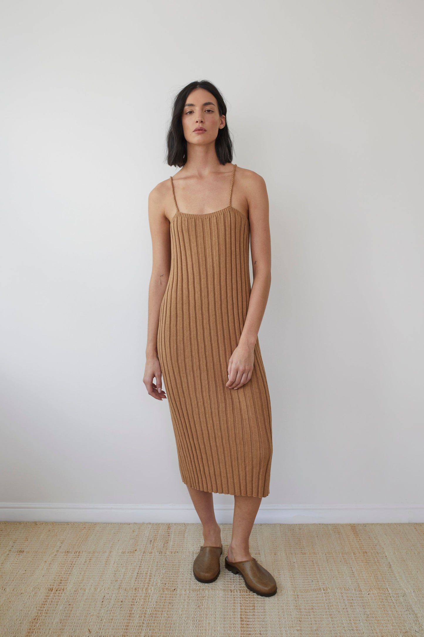 A woman with shoulder-length dark hair stands against a plain white wall, wearing the Wol Hide Rib Dress in Toffee. This mid-calf length, sleeveless knit dress features braided straps and is crafted from organic regenerative cotton. She also has on brown slip-on shoes, with her arms relaxed by her sides and a neutral expression on her face.