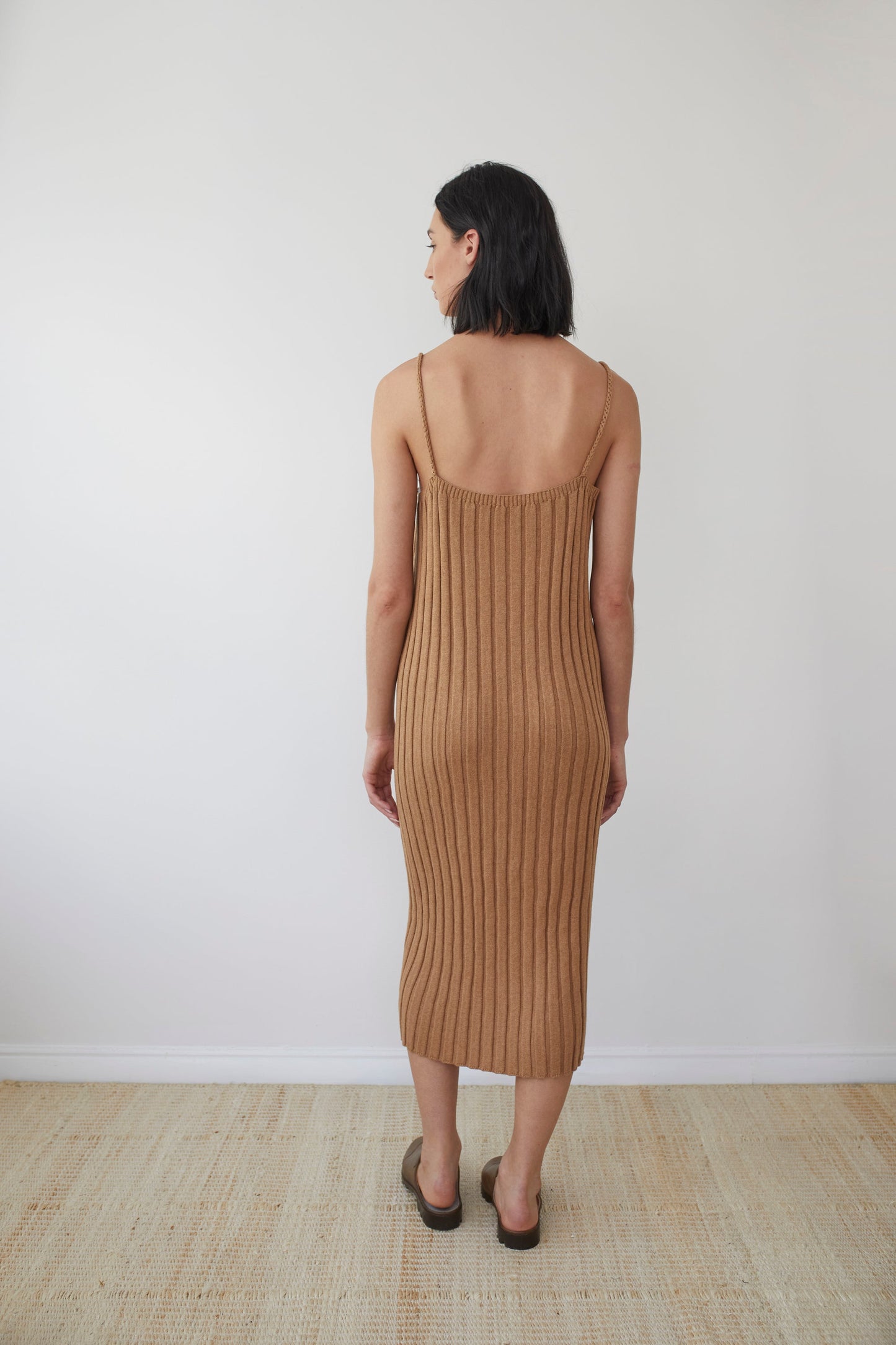 A woman with shoulder-length dark hair is standing indoors with her back to the camera. She is wearing a Wol Hide Rib Dress in Toffee, made from organic regenerative cotton, which falls below her knees, and brown flat sandals. The background features a plain white wall and a beige floor.