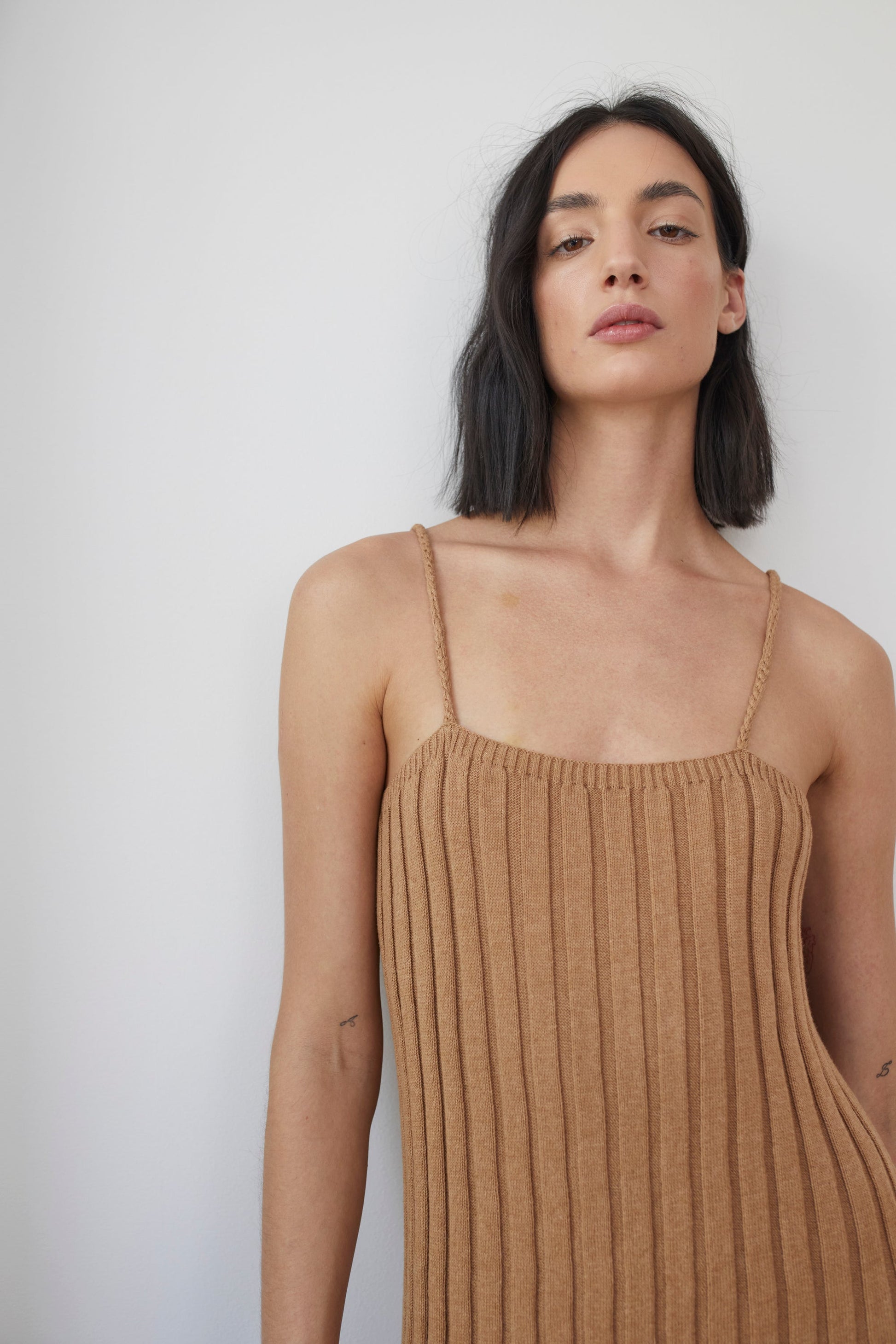 A woman with shoulder-length dark hair wears the Rib Dress in Toffee by Wol Hide, made of organic cotton and featuring thin braided straps. She stands against a plain white background, looking slightly upward with a neutral expression on her face.