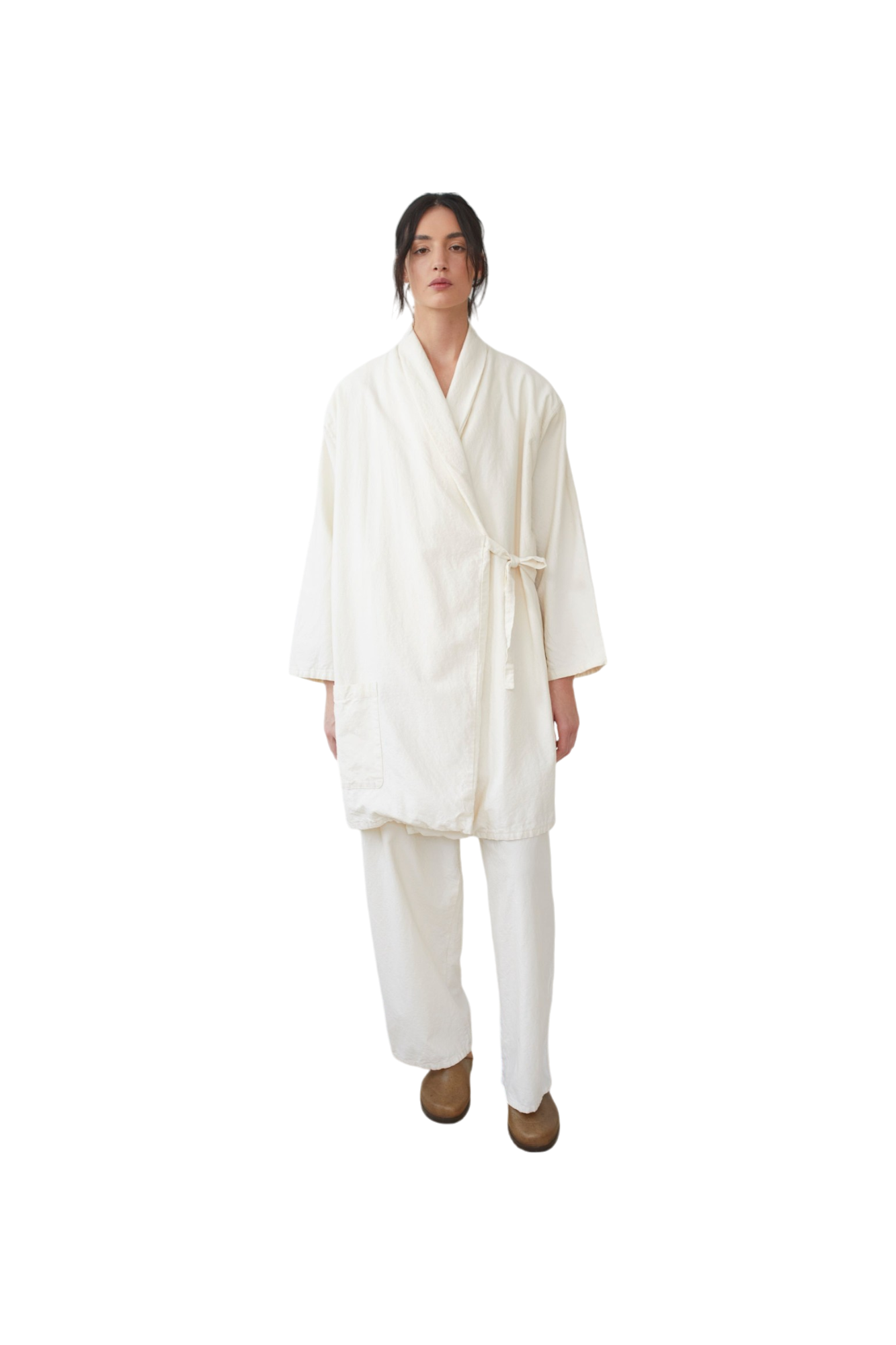 Dressed in Wol Hide's "Twill Tie Trench: Natural," the individual stands against a plain black background, showcasing a loose-fitting kimono-style outfit crafted from organic cotton. The ensemble, paired with light brown shoes, features a robe and wide-legged pants.